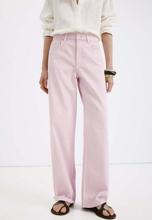 Personne portant un jean large taille haute rose clair, revers cousus, un chemisier écru, des sandales noires et plusieurs bracelets au poignet gauche.