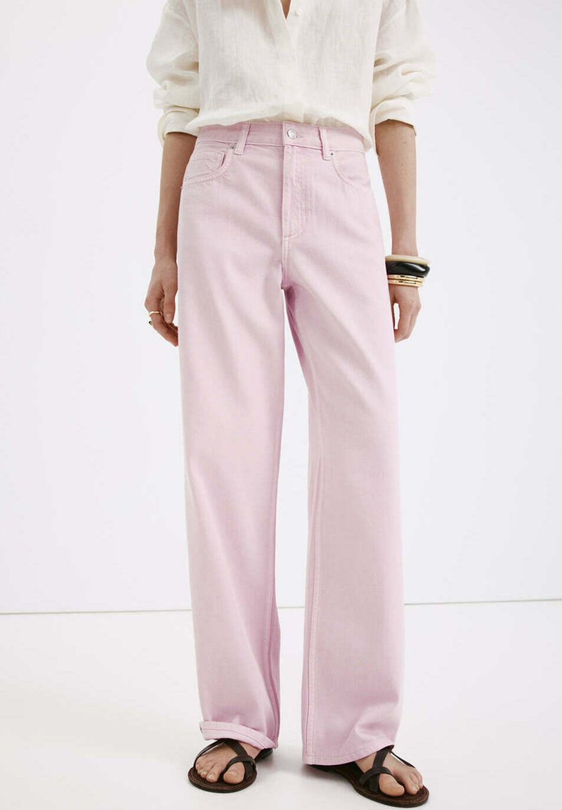 Personne portant un jean large taille haute rose clair, revers cousus, un chemisier écru, des sandales noires et plusieurs bracelets au poignet gauche.