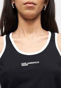 Γυναίκα που φοράει μαύρο αμάνικο τοπ με λευκή περίμετρο και κεντημένη την επιγραφή "KARL LAGERFELD JEANS" στο στήθος, με ορατά ασημένια κρίκους σκουλαρίκια.