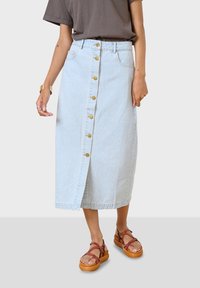 Falda midi de denim azul claro con cierre de botones delantero, bolsillos y una ligera abertura. Combinada con sandalias marrones de tiras.