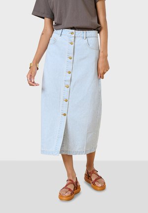 Jupe midi en denim bleu clair avec une fermeture à boutons sur le devant, des poches et une légère fente. Portée avec des sandales à lacets marron.