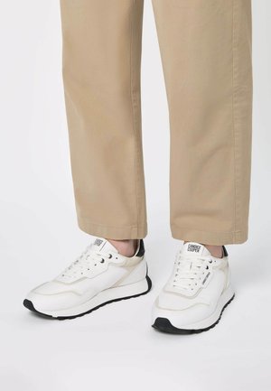 Baskets blanches avec une tige en cuir texturé, semelle en caoutchouc noir et accents beiges, associées à un pantalon beige clair.