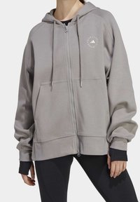 Sudadera con cremallera gris hecha de tela suave, con bolsillo frontal, capucha con cordón y un logo blanco en el pecho. Puños texturizados.