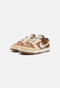 Smeđe i bež Nike tenisice s perforiranim prednjim dijelom, bijelim swoosh logom i teksturiranom gumenom potplatom, s bijelim vezicama i svijetlozelenim akcentima.