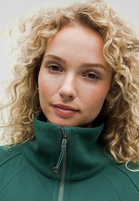 Giacca alta verde con finitura texture, dotata di una cerniera frontale e una leggera lucentezza. Capelli ricci incorniciano il viso.