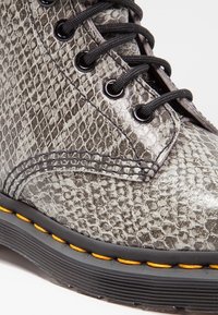 Botte en cuir gris à motif serpent, avec lacets noirs, œillets noirs, et une semelle épaisse noire avec des coutures jaunes.