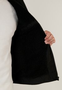 Svart jacka med en fluffig svart inredning. Har ett slätt yttertyg, hel dragkedja och ribbat avslut. Lätt strukturerad foder synligt.