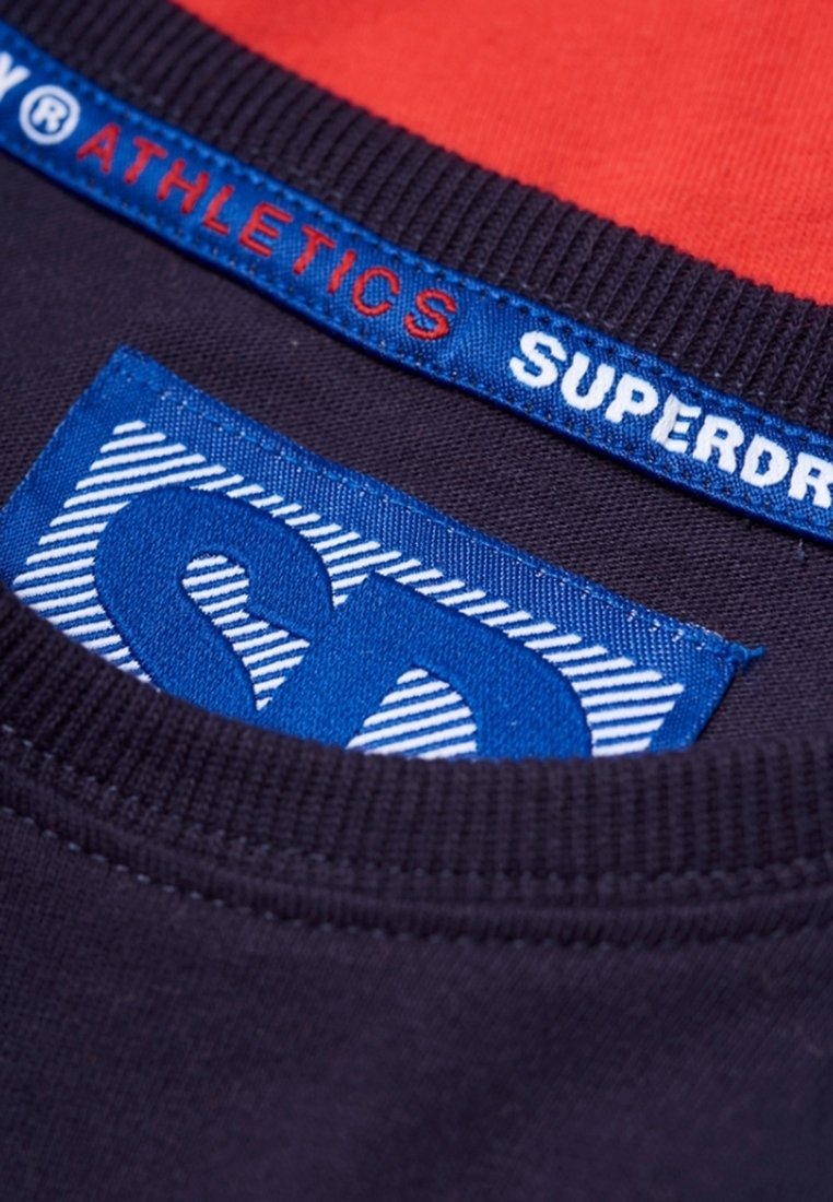 Námořnické bavlněné tričko s výstřihem, který má červenou vnitřní podšívku, modrou etiketu s bílým textem "SUPERDRY" a výraznou modrou nášivku se pruhovaným vzorem.