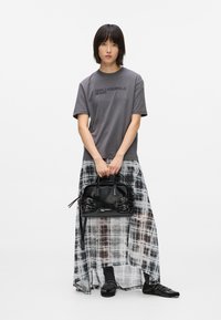 Γυναίκα στέκεται κρατώντας μια μαύρη τσάντα χεριού, φορώντας γκρι μπλουζάκι Karl Lagerfeld, διαφανή μαύρη και λευκή καρό μακριά φούστα, μαύρες κάλτσες και μαύρα παπούτσια.