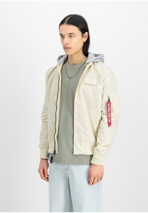 Chaqueta bomber beige claro con capucha gris, puños acanalados y cierre frontal con cremallera. Presenta un bolsillo en la manga con etiqueta roja. Llevada sobre una camiseta verde.