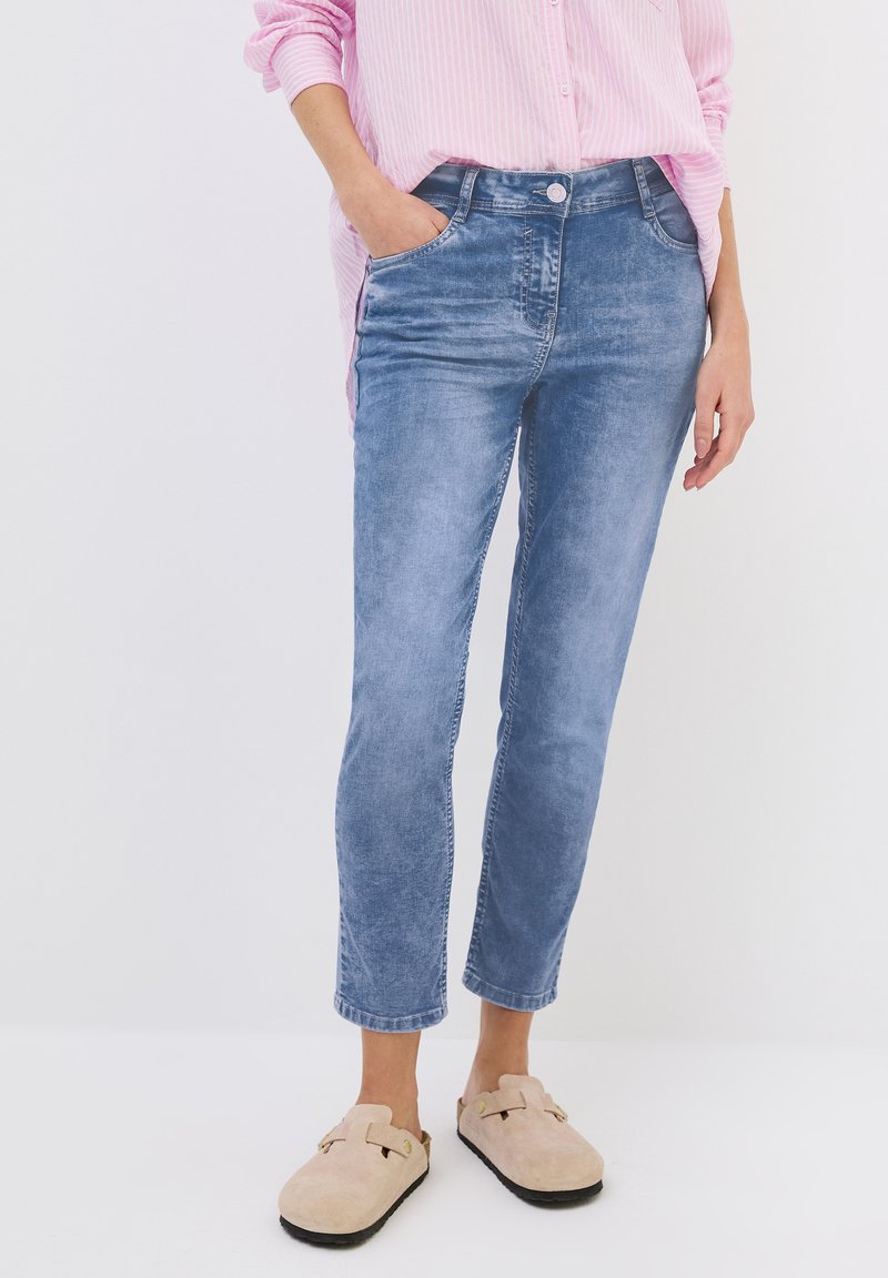 Jean en denim bleu clair avec une coupe droite, design à cinq poches et légèrement décoloré, associé à des chaussures beiges à enfiler.