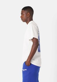 Hvid bomulds T-shirt med blå grafisk tekst på ryggen, båret med blå sweatpants med logo, set fra siden mod en neutral baggrund.