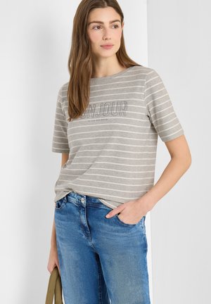 Femme portant un t-shirt à manches courtes à rayures grises et blanches avec le texte "BONJOUR", un jean bleu, tenant un sac vert olive, debout devant un fond blanc.