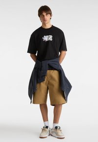 Black graphic T-shirt with "VANS" logo, khaki shorts, navy button-up tied around the waist, paired with cream and green sneakers.