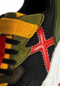 Zapatilla multicolor con tejido negro, detalles en ante amarillo y verde, costuras rojas y elementos texturizados sobre una suela blanca.