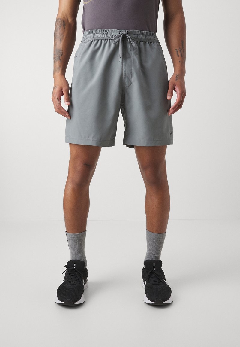 Athlète portant un short de sport gris avec cordon, un haut sans manches sombre, des chaussettes grises et des chaussures de course noires, debout sur un fond uni.