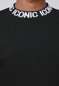 Zwart geribbeld sweater met een ronde halslijn en witte gebreide tekst "ICONIC" op de kraag. Gladde textuur en nauwsluitend ontwerp.
