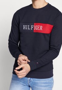 Muž v námořnickém modrém svetru s vodorovnými červenými a námořnickými pruhy a nápisem "HILFIGER", doplněný o světle modré džíny.