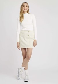 Haut blanc à manches longues, mini-jupe en tweed jaune clair avec deux boutons dorés, et baskets blanches avec des chaussettes blanches jusqu'à la cheville. Fond uni.
