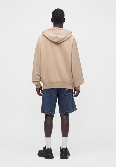 Personne debout de dos, portant un sweat à capuche beige, un short en jean bleu, des chaussettes blanches et des baskets noires sur un fond uni.