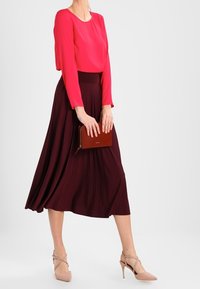Rode sheer blouse met lange mouwen gecombineerd met een donker burgundy geplooide midi rok. Beige hoge hakken en een bruine clutch tas maken de look compleet.