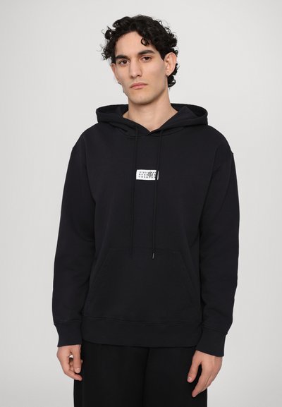 Hoodie en coton noir avec une poche kangourou, capuche à cordon de serrage et une petite étiquette graphique blanche sur la poitrine. Présente des poignets et un ourlet côtelés.