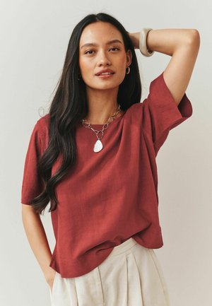 Hosszú sötét hajú nő, aki rozsdabarna rövid ujjú felsőt, bézs nadrágot, vastag medált, aranykarika fülbevalókat és bézs karkötőt visel.