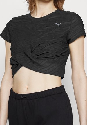 Kobieta w czarnej teksturowanej skróconej koszulce Puma skręconej w talii i czarnych spodniach dresowych z elastycznym pasem.