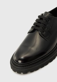 Chaussure en cuir noir avec une finition brillante, bout rond et semelle en caoutchouc texturée. Présente des lacets noirs et un logo en relief sur le côté.