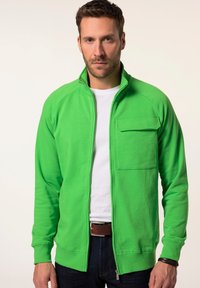 Zelená zip-up mikina vyrobená z měkké tkaniny, s přední kapsou, žebrovanými manžetami a stojáčkem, nošená přes bílý tričko.