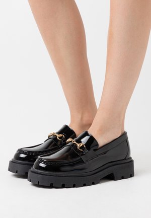 Mocassins en cuir verni noir avec des semelles en caoutchouc épaisses et un détail en biton doré portés pieds nus sur un fond blanc.