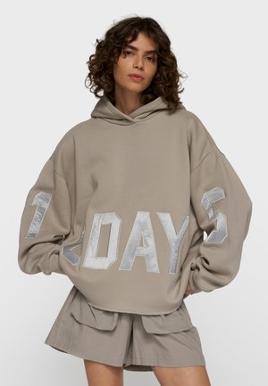 Kvinde med krøllet hår iført oversized beige hættetrøje med sølvfarvet "1DAY5" tekst og beige shorts, stående foran en ensfarvet baggrund.