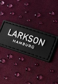 Țesătură maro cu picături lucioase, cu o suprafață texturată. O pată dreptunghiulară neagră care prezintă marca "LARKSON HAMBURG" cu litere albe.