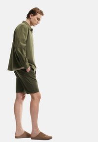 Olivenfarvet skjorte med lange ærmer og knapdetaljer, parret med olivenfarvede shorts og brune slip-on sko. Modellen står sidelæns.