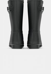 Bottes de pluie en caoutchouc noir avec des côtés texturés, des sangles ajustables et des semelles renforcées, présentant des rainures verticales et une finition lisse.