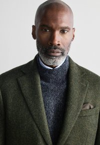 Dunkelgrüner Tweed-Blazer über einem grauen Strickpullover, mit einem hellblauen Kragenhemd darunter. Ein graues Einstecktuch vervollständigt den Look.