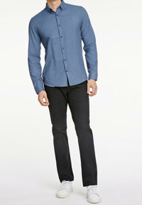 Camicia azzurra a maniche lunghe con bottoni scuri, di texture liscia, abbinata a pantaloni neri e sneakers bianche, in piedi contro uno sfondo neutro.