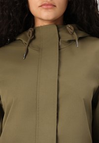 Giacca impermeabile verde oliva con colletto alto, chiusure a toggle e una superficie liscia. Presenta un pannello centrale con zip e cuciture minime.