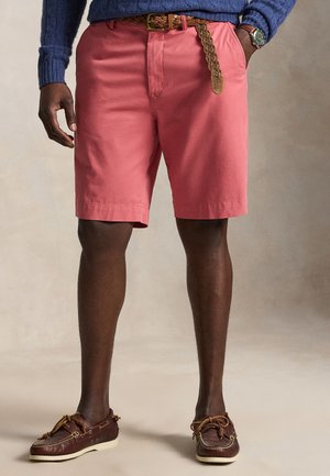 Mand iført koralrosa shorts med et flettet brunt bælte, blå sweater, brune læder bådsko og et armbåndsur, stående med hænderne i lommerne.