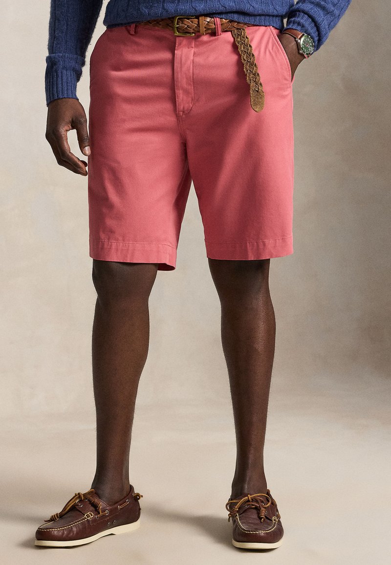 Mann trägt korallrosa Shorts mit geflochtenem braunem Gürtel, blauen Pullover, braune Leder-Bootsschuhe und eine Armbanduhr, steht mit den Händen in den Taschen.