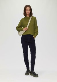 Grüner gerippter Pullover, marineblaue Cordschnurrhose und schwarze klobige Stiefel. Beiger Umhängetasche. Modell steht vor einem neutralen Hintergrund.