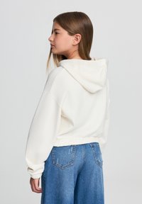 Felpa bianca con cappuccio, dal design cropped, orlo a coste e maniche oversized, abbinata a jeans a gamba larga blu.