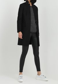 Cappotto nero in misto lana con taglio dritto, chiusura con un solo bottone e tasche laterali, abbinato a leggings neri in ecopelle e sneakers bianche.