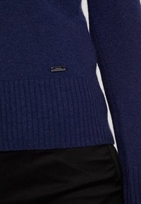 Maglione a maglia blu navy con orlo a coste, caratterizzato da una piccola etichetta metallica del marchio. Realizzato in materiale morbido con una finitura testurizzata.