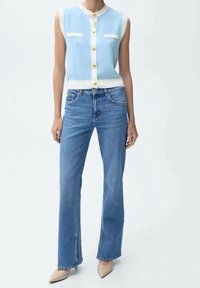 Top de punto sin mangas de color azul claro con acentos blancos, botones dorados y dos bolsillos frontales, combinado con jeans de tiro alto azules y tacones beige.