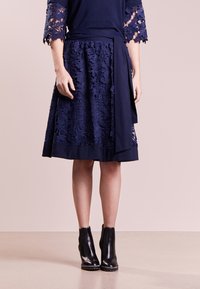 Laivansinis mekko, jossa on kukkakuvioinen pitsimuotoilu, kapea vyötärö, laskostettu hame, jossa on kiinteä helma, yhdistetty mustiin nilkkasaappaisiin.