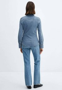 Chemise en denim bleu clair à manches longues, poignets boutonnés et empiècement sur le devant ; associée à un jean évasé bleu et des chaussures noires.