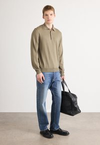 Ung mann iført beige langermet polo, blå jeans, svarte sko, holder en svart skinn duffelbag, står på grått gulv med hvit vegg i bakgrunnen.