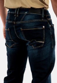 Jean en denim bleu foncé avec une coupe ajustée, comportant deux poches arrière, une couture contrastante et une ceinture noire texturée.