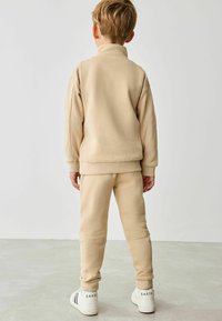 Beige fleece træningsdragt med høj krave og ribbede manchetter, parret med hvide sneakers med sort skrift på hælen.